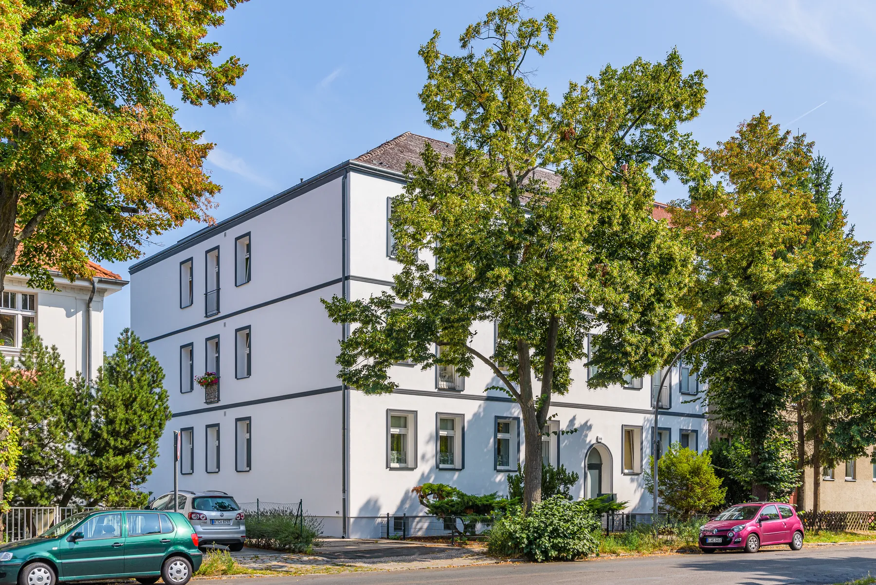 Weiß verputztes Mehrfamilienhaus hinter hohen Bäumen