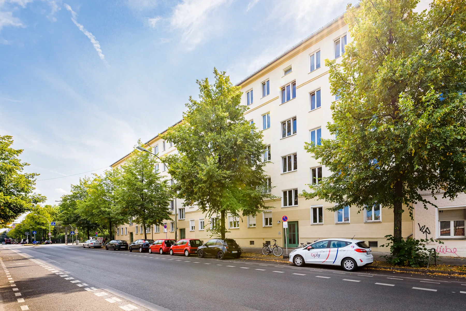 Straße mit hellem Mehrfamilienhaus und Bäumen im Sonnenlicht