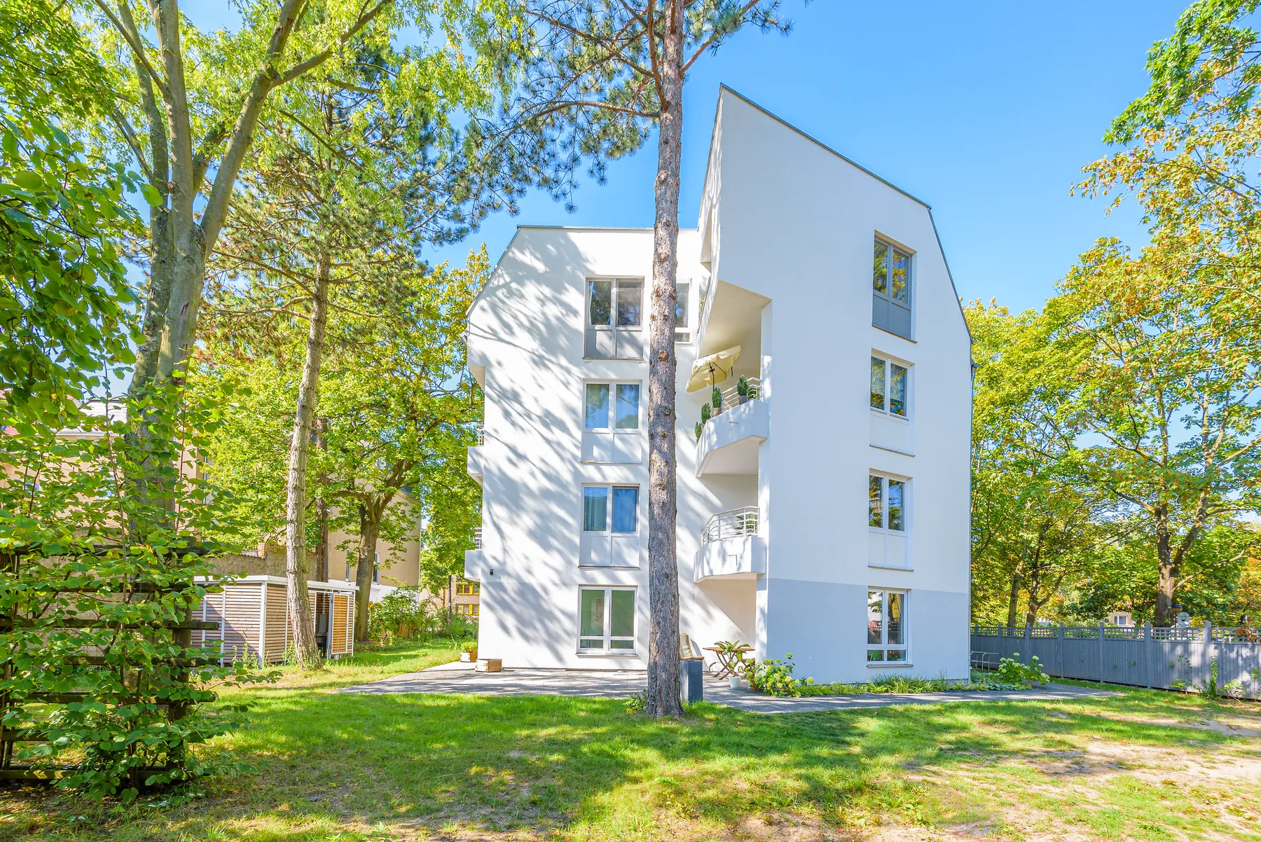 Weißes Mehrfamilienhaus mit Balkonen zwischen hohen Bäumen